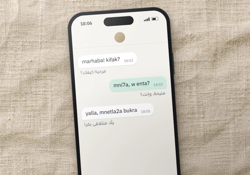 Smartphone on linen showing a Levantine chat in Arabizi, “marhaba! kifak?”, with the Arabic-script version under each bubble