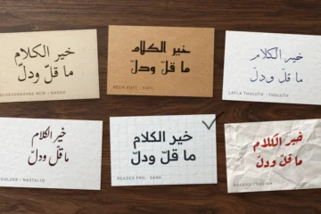 Six cards on a walnut desk, each with the proverb خير الكلام ما قلّ ودلّ in a different Arabic font style, one ticked in pencil