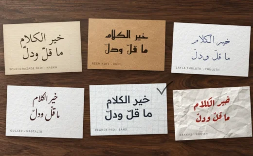 Six cards on a walnut desk, each with the proverb خير الكلام ما قلّ ودلّ in a different Arabic font style, one ticked in pencil